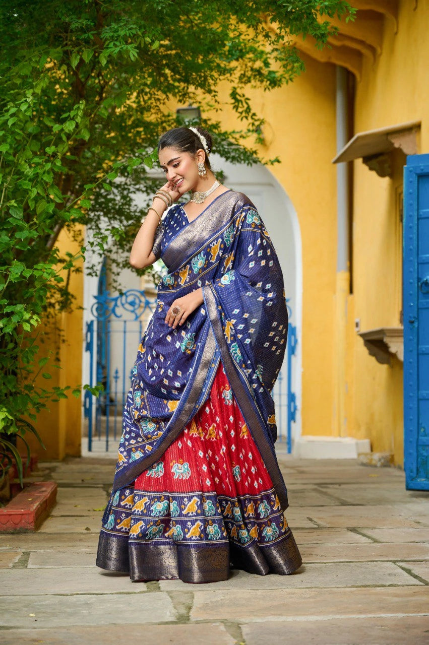 Woman in a blue and red saree standing in front of a yellow wall with greenery.